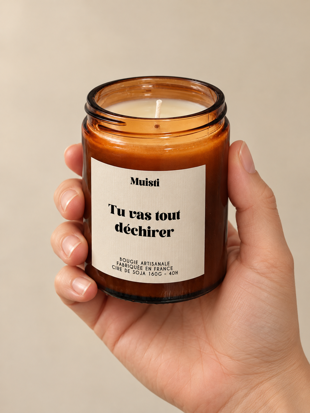 Bougie Muisti « Tu vas tout déchirer » en pot ambré, tenue dans une main sur fond beige.