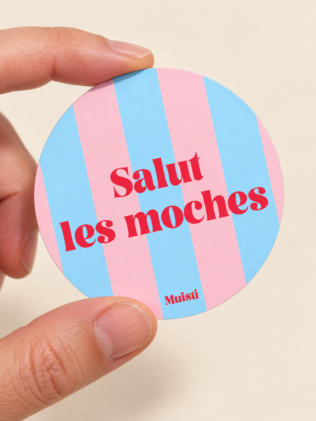 Sticker Muisti Salut les moches tenu entre le pouce et l’index sur fond ivoire