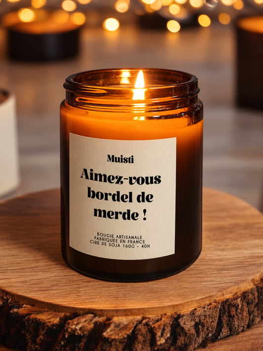 Bougie Muisti « Aimez-vous bordel de merde » allumée, en pot ambré sur un socle en bois.