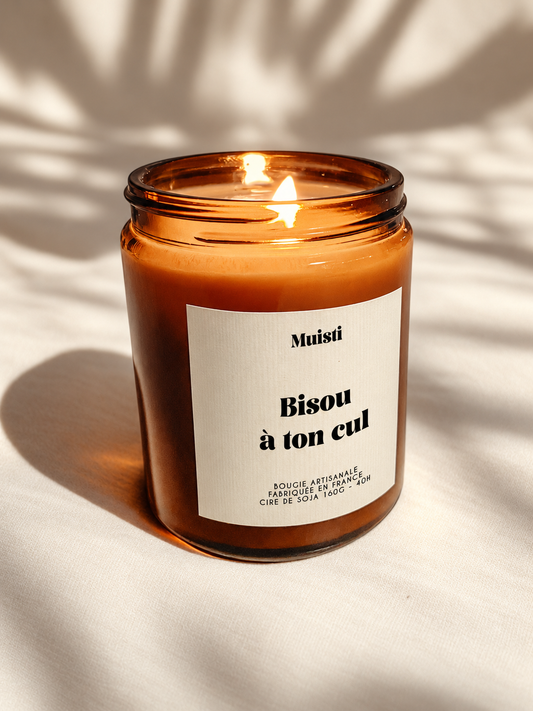 Bougie Muisti « Bisou à ton cul » allumée, en pot ambré sur un tissu ivoire.