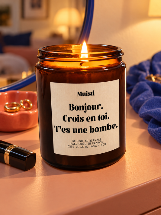 Bougie Muisti « Bonjour. Crois en toi. T’es une bombe. » allumée sur une coiffeuse corail, en soirée.