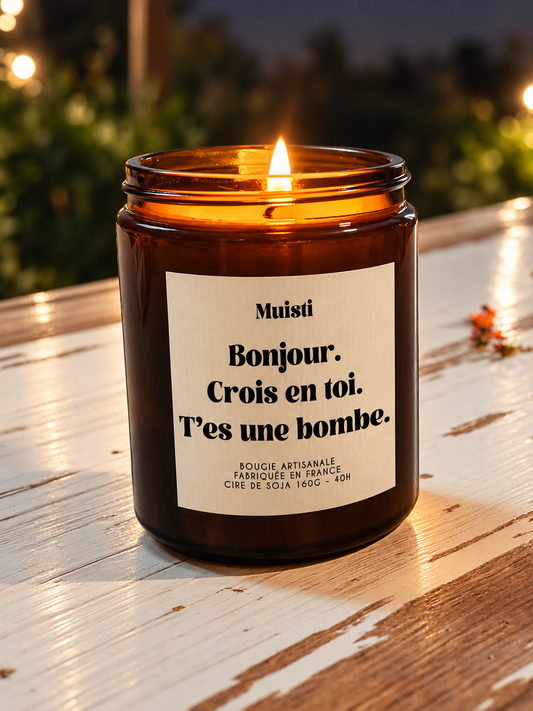 Bougie Muisti « Bonjour. Crois en toi. T’es une bombe. » allumée, en pot ambré sur une table en bois patiné.