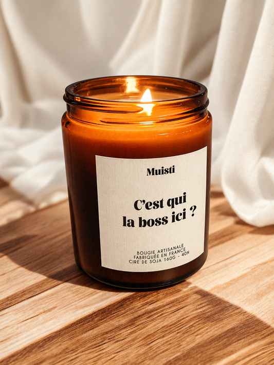 Bougie Muisti « C’est qui la boss ici ? » allumée, en pot ambré sur une table en bois devant un tissu ivoire.
