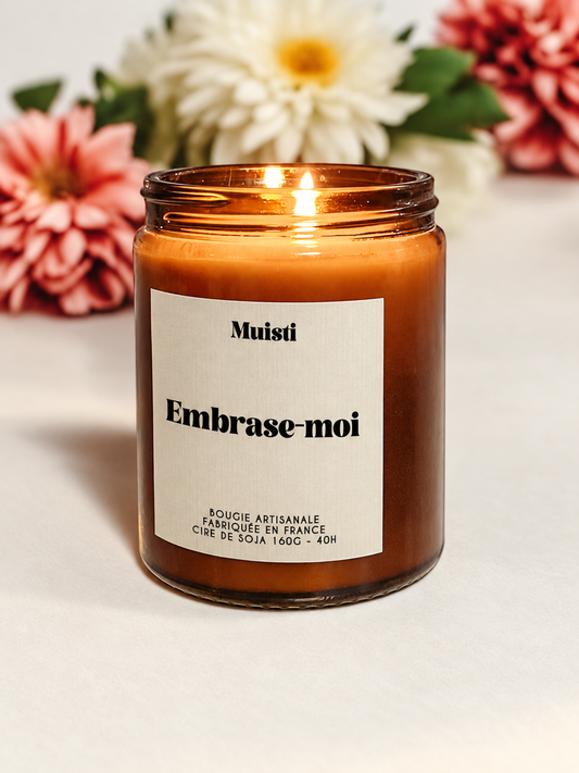 Bougie Muisti « Embrase-moi » allumée, en pot ambré devant des fleurs.