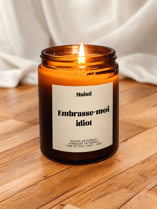 Bougie Muisti « Embrasse-moi idiot » allumée, en pot ambré sur une table en bois.