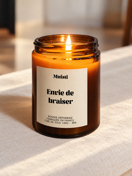 Bougie Muisti « Envie de braiser » allumée, en pot ambré sur un tissu ivoire, dans une lumière naturelle chaleureuse.