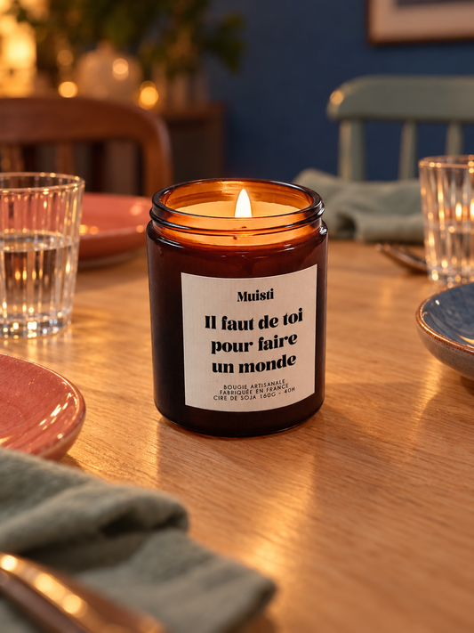 Bougie Muisti « Il faut de toi pour faire un monde » allumée sur une table de dîner avec de la vaisselle colorée.