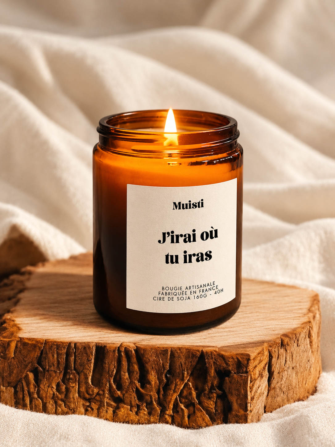 Bougie Muisti « J’irai où tu iras » allumée, en pot ambré sur un socle en bois.
