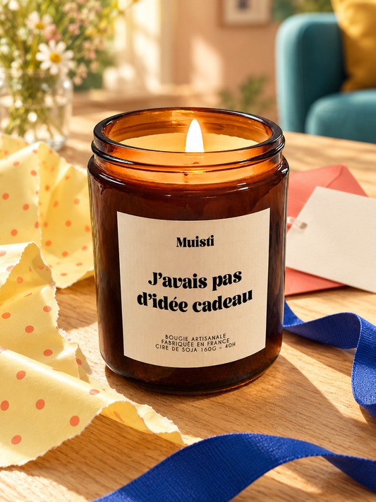 Bougie Muisti « J’avais pas d’idée cadeau » allumée, avec papier cadeau jaune et ruban bleu.
