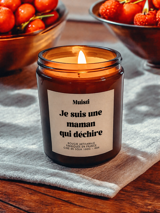 Bougie Muisti « Je suis une maman qui déchire » allumée, en pot ambré sur une table en bois et un tissu clair.