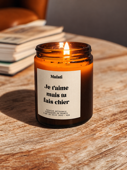 Bougie Muisti « Je t’aime mais tu fais chier » allumée, en pot ambré dans son décor d’origine.