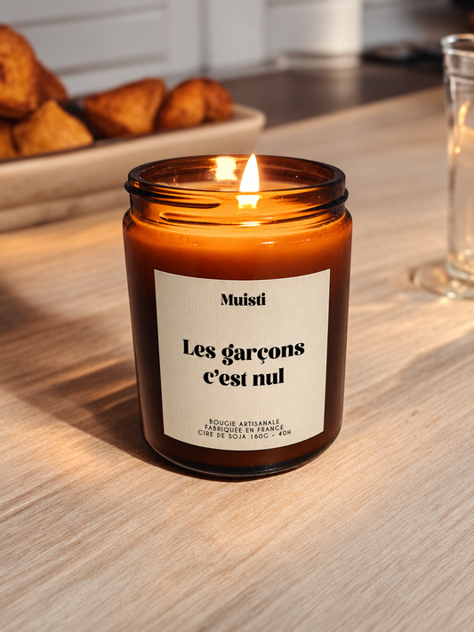 Bougie Muisti « Les garçons c’est nul » allumée, en pot ambré sur une table en bois, avec des croissants en arrière-plan.
