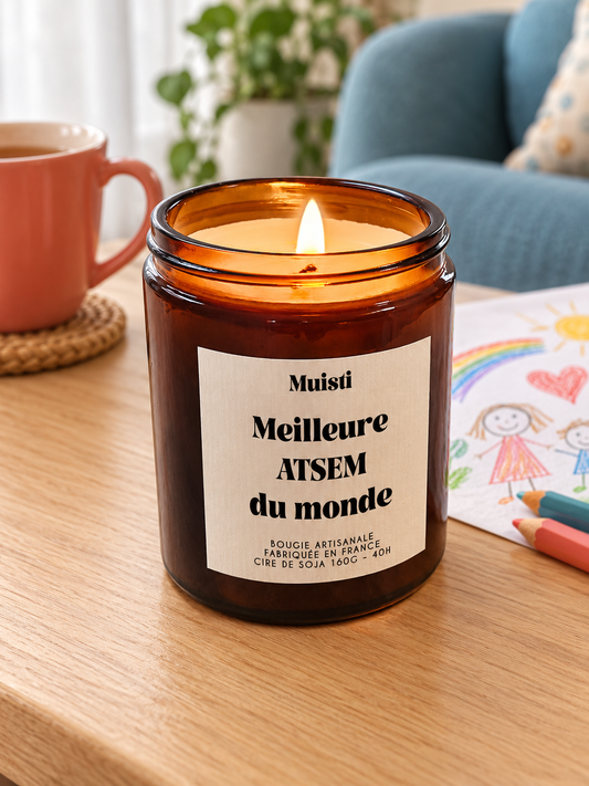 Bougie Muisti « Meilleure ATSEM du monde » 160 g allumée sur une table dans un intérieur à la lumière douce du jour.