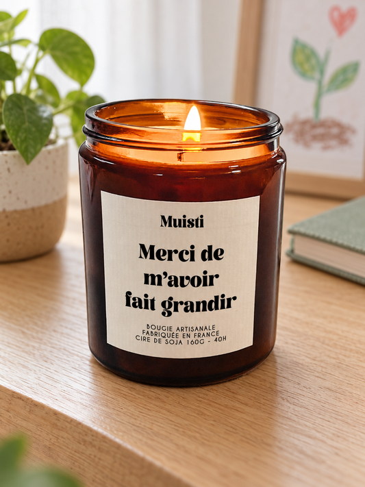 Bougie Muisti « Merci de m'avoir fait grandir » 160 g allumée sur une console en bois, près d’une plante et d’un dessin d’enfant.