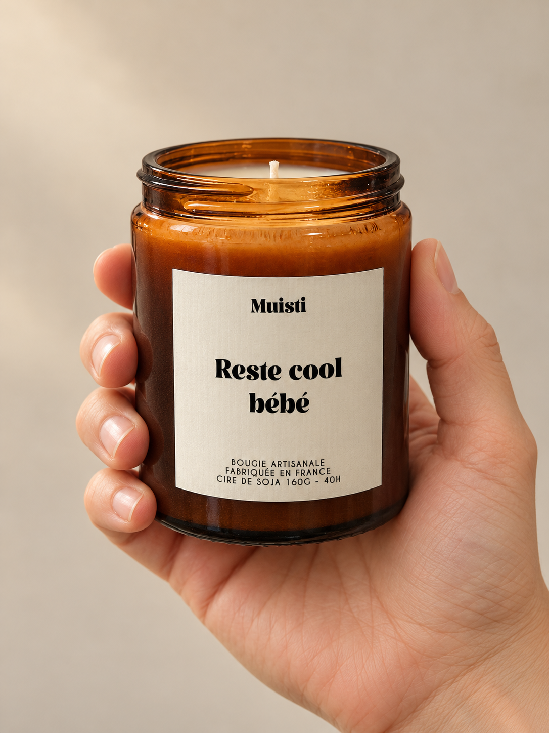 Bougie Muisti « Reste cool bébé » en pot ambré, tenue dans une main sur fond beige.