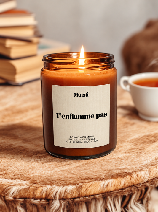 Bougie Muisti « T’enflamme pas » allumée, en pot ambré sur une table en bois près de livres et d’une tasse.