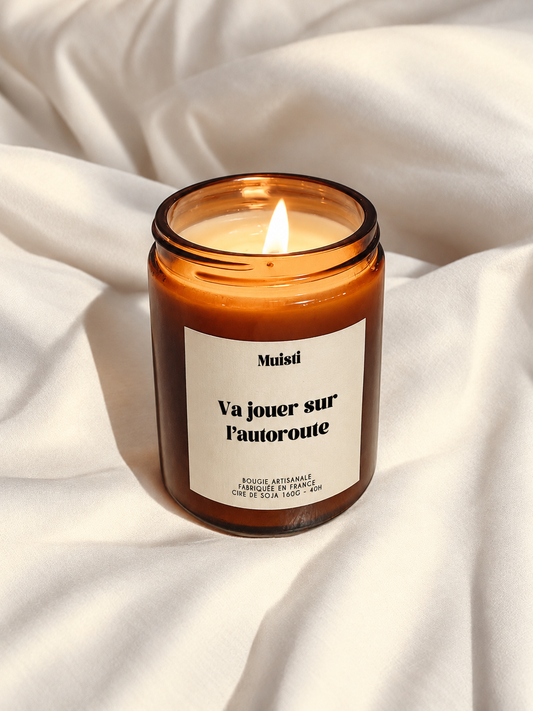 Bougie Muisti « Va jouer sur l’autoroute » allumée, en pot ambré sur un tissu ivoire.