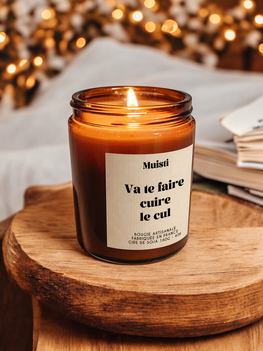 Bougie Muisti « Va te faire cuire le cul » allumée, en pot ambré sur un socle en bois.