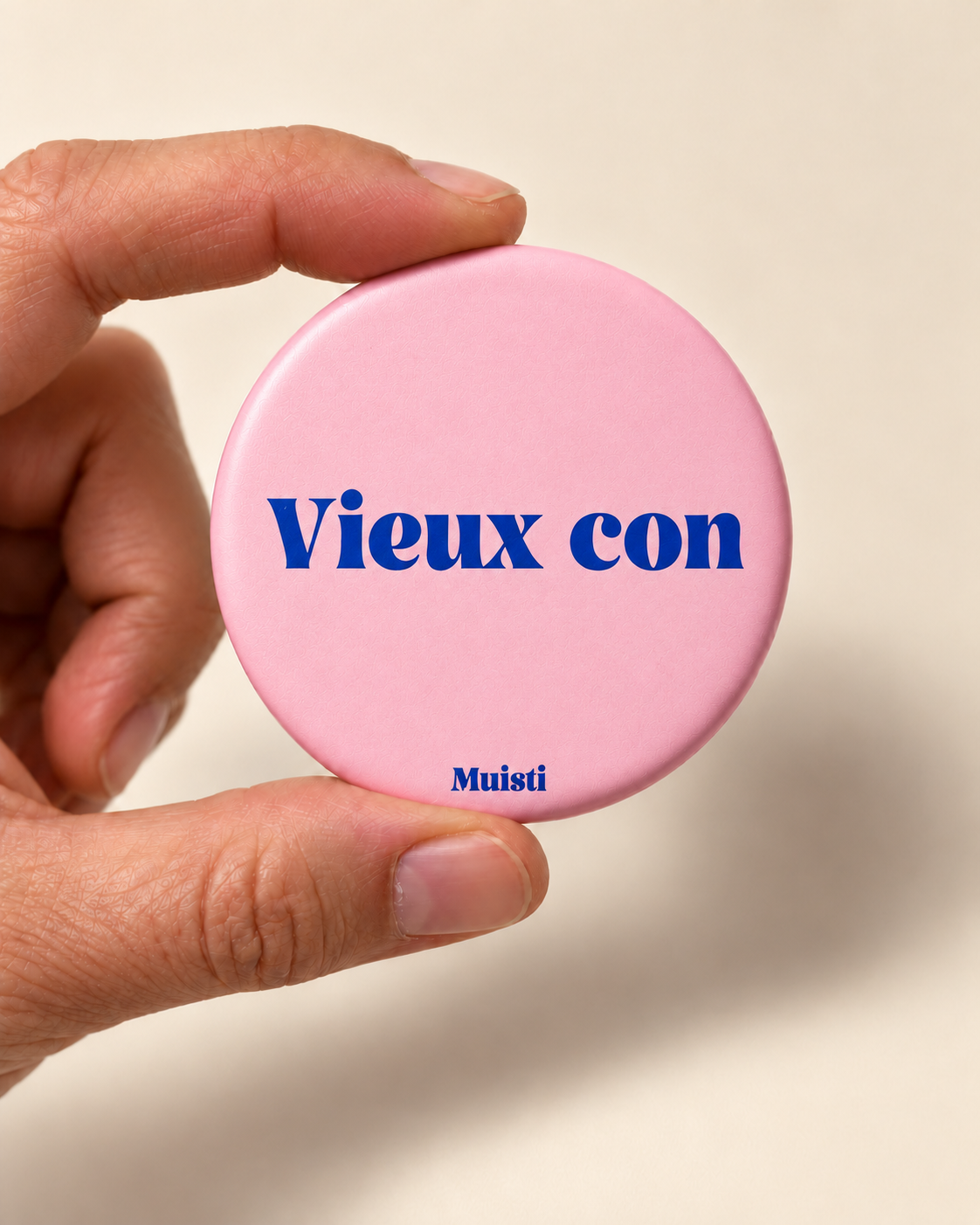 Magnet Muisti « Vieux con » tenu entre le pouce et l’index sur fond ivoire.