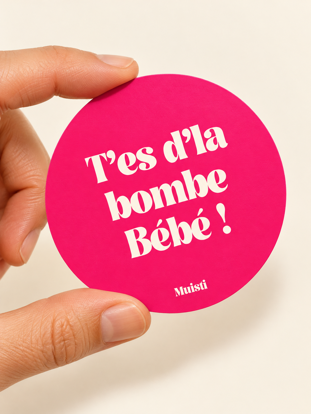 Sticker Muisti T’es d’la bombe Bébé ! tenu entre le pouce et l’index sur fond ivoire