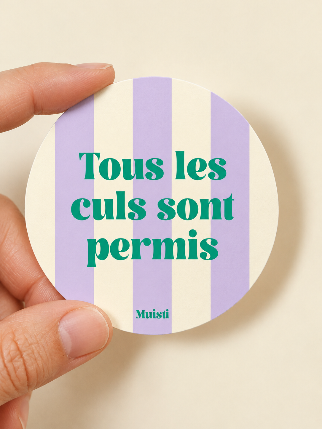 Sticker Muisti Tous les culs sont permis tenu entre le pouce et l’index sur fond ivoire
